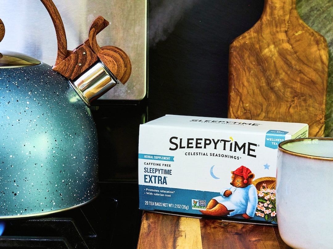 A box of SleepyTime Caffeine Free SleepyTime Extra herbal tea sits on a wooden surface next to a speckled teal kettle and a white mug with a gold rim.