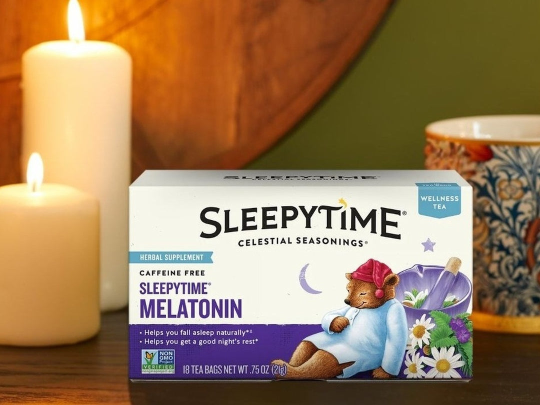 A box of Sleepytime Melatonin herbal supplement tea by Celestial Seasonings, styled on a wooden surface with lit white candles and a patterned ceramic mug in the background.